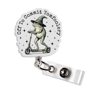 May include: A white badge reel with a whimsical illustration of a frog wearing a witch hat and riding a scooter. The text "Off To Commit Tomfoolery" is printed around the edge of the badge. The reel has a clear plastic strap and a metal clip.