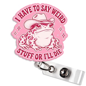 May include: A pink badge reel featuring a circular design with a frog wearing a cowboy hat. The text I HAVE TO SAY WEIRD STUFF OR I'LL DIE is printed in dark pink capital letters around the frog. The badge reel has a clear plastic strap.