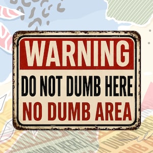 May include: A rectangular metal sign with a distressed, weathered finish. The sign features a red top banner with the word WARNING in white, followed by the black text DO NOT DUMB HERE and the red text NO DUMB AREA on a cream background.