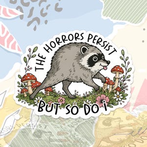 May include: A die-cut sticker featuring a grey raccoon walking through a patch of green grass, red and white mushrooms, and pink flowers. The text THE HORRORS PERSIST BUT SO DO I is printed in black capital letters around the raccoon illustration.