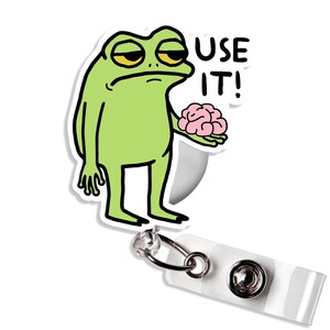 May include: A green frog cartoon character with yellow eyes and a sad expression, holding a pink brain. The text "USE IT!" is printed in black. The frog is on a white retractable badge reel.