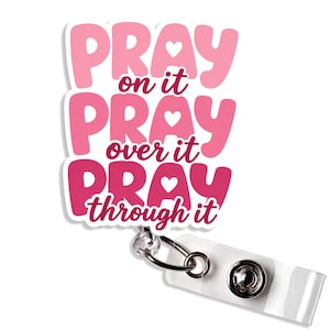 May include: A badge reel featuring a die-cut topper with the text PRAY on it, PRAY over it, PRAY through it in shades of pink and dark magenta. Each word PRAY includes a small white heart. The topper is attached to a clear plastic strap and clip.