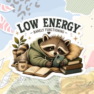 May include: A die-cut sticker featuring a raccoon wearing a green hoodie, lying down while looking at a smartphone. The text reads "LOW ENERGY" and "BARELY FUNCTIONING". The raccoon is surrounded by a coffee mug, an open book, and a stack of books.