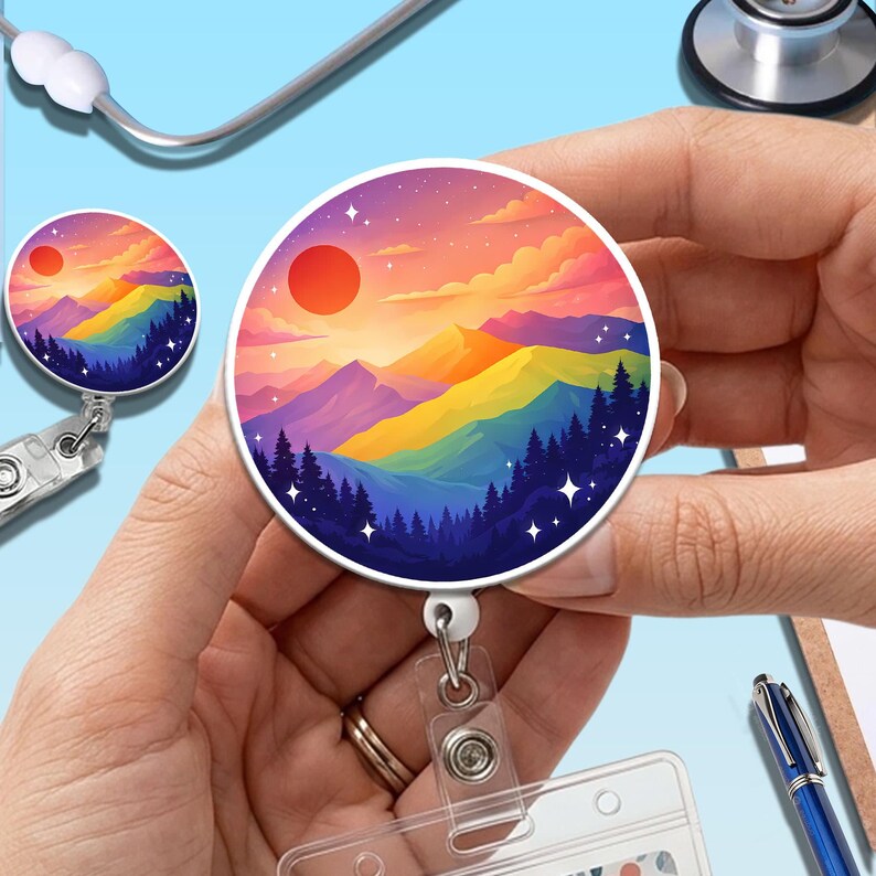 May include: A round badge reel featuring a colorful mountain landscape with a red sun, rainbow-colored mountains, and dark green trees. The badge reel is held in a person's hands, with a clear plastic ID holder attached.