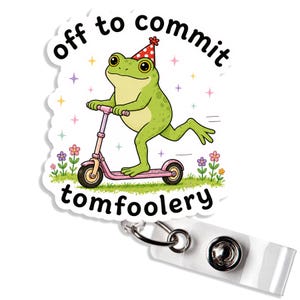 May include: A badge reel featuring a green frog wearing a red polka-dot party hat, riding a pink scooter. The text reads "off to commit tomfoolery" with small colorful flowers and stars in the background on a white die-cut sticker shape.