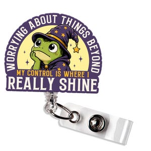 May include: A badge reel featuring a green frog wearing a purple wizard hat with yellow stars. The text reads: WORRYING ABOUT THINGS BEYOND MY CONTROL IS WHERE I REALLY SHINE. The design is circular with a clear plastic strap and a metal snap button.