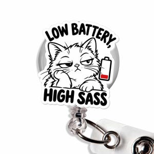 May include: A circular badge reel featuring a black and white illustration of a cat with a tired expression, a low battery icon with a red bar, and the text LOW BATTERY, HIGH SASS printed in bold black capital letters.