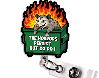 May include: A badge reel with a cartoon opossum in a green dumpster surrounded by flames. The text on the dumpster reads "THE HORRORS PERSIST BUT SO DO I". The reel has a clear plastic strap and a metal clip.