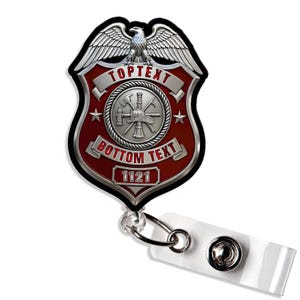 May include: A red and silver badge-shaped retractable ID badge reel featuring an eagle emblem at the top, a central circular firefighter tool icon, and the text TOPTEXT, BOTTOM TEXT, and 1121. A clear plastic strap with a metal snap is attached.