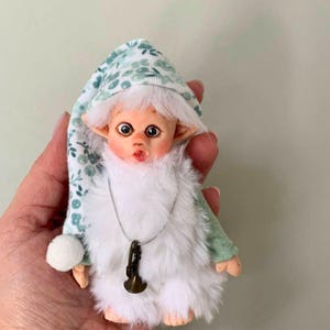Könnte beinhalten: Eine handgefertigte Puppe mit einem skurrilen Design. Die Puppe hat einen hellen Teint, große Augen und einen überraschten Gesichtsausdruck. Sie trägt eine grün-weiß gemusterte Mütze, einen flauschigen, weißen, fellartigen Körper und einen kleinen Anhänger in Form eines Messinghorns.