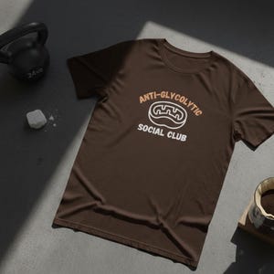 Op de afbeelding: Een bruin t-shirt met de tekst "ANTI-GLYCOLYTIC SOCIAL CLUB" in wit en oranje. Een zwarte kettlebell, gemarkeerd met 24 kg, en een klein wit object op de achtergrond.