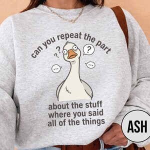 Puede incluir: Sudadera gris claro con un dise&ntilde;o de ganso de dibujos animados y el texto "can you repeat the part about the stuff where you said all of the things." La palabra "ASH" est&aacute; en un c&iacute;rculo negro.