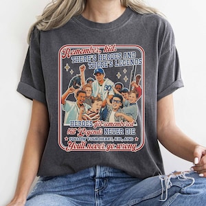 May include: Dark gray t-shirt featuring a vintage-style graphic with the text "Remember kid, There's Heroes and There's Legends." The graphic includes illustrations of young baseball players and the phrase "Heroes get remembered but Legends Never Die."