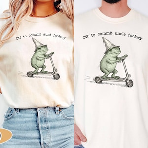 Puede incluir: Camisetas color crema con una ilustración de rana. La rana, con un sombrero de mago, monta un patinete. Una camiseta dice "Off to commit aunt foolery", la otra dice "Off to commit uncle foolery."