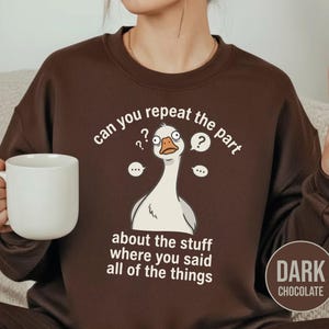 Puede incluir: Sudadera marr&oacute;n chocolate oscuro con un gr&aacute;fico blanco de un ganso confundido y el texto "can you repeat the part about the stuff where you said all of the things". Una taza blanca se sostiene en la mano.