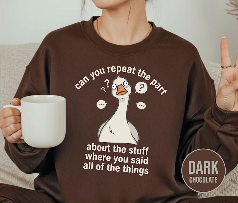 Puede incluir: Sudadera marr&oacute;n chocolate oscuro con un gr&aacute;fico blanco de un ganso confundido y el texto "can you repeat the part about the stuff where you said all of the things". Una taza blanca se sostiene en la mano.
