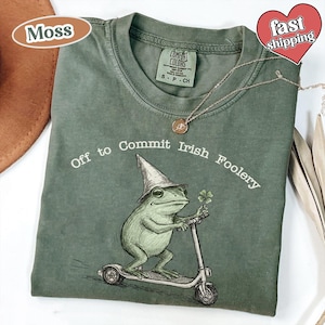 以下が含まれることがあります： モスグリーンのTシャツ。テキスト「Off to Commit Irish Foolery」と、帽子をかぶり四つ葉のクローバーを持った、スクーターに乗るカエルのグラフィック。金のネックレスがシャツの上にかけられています。