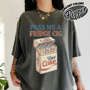 May include: Dark grey t-shirt with the text "PASS ME A FRIDGE CIG" in blue and red. Below the text is a graphic of a Diet Coke carton with six cans. The shirt also has the text "COMFORT COLORS Pepper".