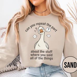 Puede incluir: Sudadera de color arena con una ilustraci&oacute;n de un ganso de dibujos animados y el texto "can you repeat the part about the stuff where you said all of the things." La palabra "SAND" est&aacute; en un c&iacute;rculo en la parte inferior derecha.