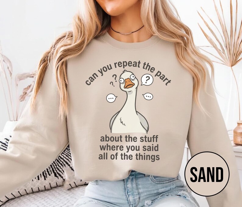 Puede incluir: Sudadera de color arena con una ilustraci&oacute;n de un ganso de dibujos animados y el texto "can you repeat the part about the stuff where you said all of the things." La palabra "SAND" est&aacute; en un c&iacute;rculo en la parte inferior derecha.
