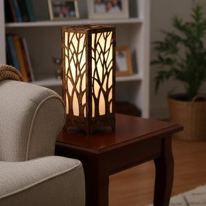 May include: A rectangular table lamp with a dark brown wooden frame featuring a laser-cut tree branch pattern. The lamp emits a warm, soft glow through a translucent white inner panel, creating a silhouette effect of forest branches.