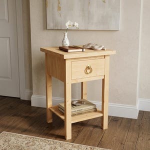 May include: A light-colored wooden nightstand with a single drawer and a lower shelf. The drawer has a gold-colored ring pull. A small vase with flowers, a notebook, and a sleep mask rest on top. Books with the text "DESIGN DESIGN" are on the shelf.