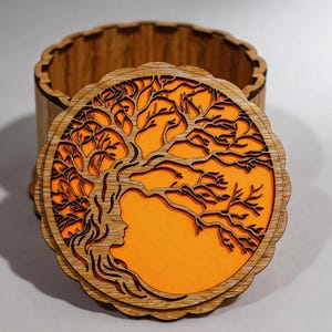 May include: A round wooden box with a tree of life design on the lid. The lid has a layered design with a tree silhouette and an orange background. The box is made of light brown wood and has a scalloped edge.