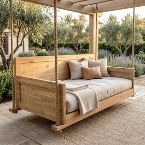May include: A wooden swing bed with a light beige mattress and pillows. The bed is suspended by thick rope from a wooden pergola. A beige blanket is folded on the bed. The background features a garden with trees and lavender bushes.