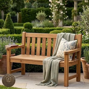 Può includere: Una panchina da giardino in legno con schienale e seduta a doghe, drappeggiata con una coperta verde salvia e un cuscino decorativo con motivo floreale blu e bianco. Un paio di occhiali poggia su un libro sul bracciolo sinistro.