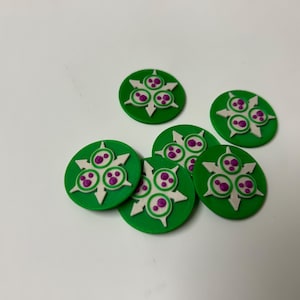 May include: A collection of six green, circular game tokens. Each token features a white starburst design with three purple circles and white accents. The tokens are scattered on a white surface.