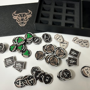 May include: Black game token set with various designs and text, including "+1 HIT", "DMG", and "Ward 2+". A black box with a bull logo and a compartmented tray is also present.