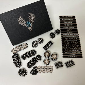 May include: A black box with a deer emblem and various game tokens. The tokens include round and rectangular pieces with text and symbols, such as "Hit", "Ward", and "First Strike". The game pieces are in black, white, and tan.
