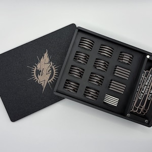 May include: A black rectangular storage box with a matching lid featuring a gold lightning bolt emblem. The interior contains twelve circular tokens with silver and black stripes, plus a side compartment holding several thin rectangular markers.