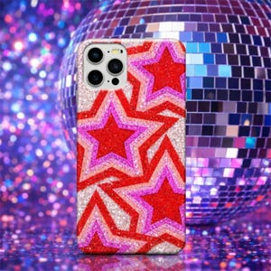 May include: A phone case with a glittery design featuring layered red, pink, and white stars. The case has a camera cutout and a shiny, textured surface. The background includes a disco ball and sparkling purple lights.