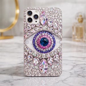 Könnte beinhalten: Funkelnde Handyhülle mit Kristall-Design. Die Hülle zeigt ein großes Augendesign mit blauen, rosa und lila Akzenten. Die Hülle ist mit kleinen, klaren Kristallen und größeren, tropfenförmigen Kristallen bedeckt.