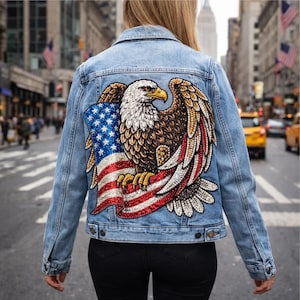 May include: Light blue denim jacket featuring a large, embellished American flag and a detailed eagle design on the back. The eagle has a gold beak and is clutching the flag. The jacket has a classic button-down design.