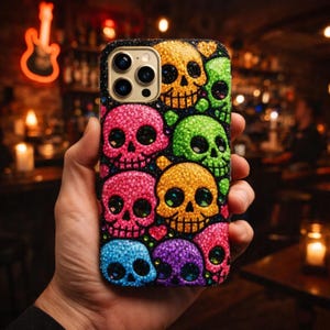 Puede incluir: Una funda de teléfono colorida con un patrón de calaveras brillantes en varios colores, incluyendo rosa, verde, amarillo, azul y morado. La funda tiene un borde negro y un recorte para la cámara.