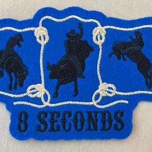May include: A blue felt patch with a rodeo theme. The patch features three black silhouettes of a bucking horse, a bull rider, and a bronco rider, framed by a white rope design. The text "8 SECONDS" is embroidered in black.