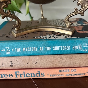 Peut inclure: Une pile de trois livres anciens, dont les titres incluent "The Mystery at the Shuttered Hotel" et "Three Friends". Les livres sont de couleurs bleu, pêche et beige, avec des accents dorés. Un objet décoratif en laiton est posé sur les livres.