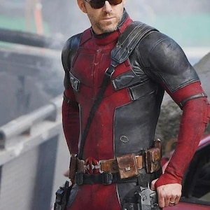 Puede incluir: Una persona con un traje t&aacute;ctico rojo y negro de acabado texturizado, un cintur&oacute;n utilitario de cuero marr&oacute;n con hebilla circular y fundas negras con pistolas. El conjunto incluye gafas de sol oscuras y una correa al hombro.