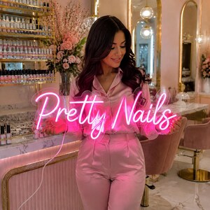 May include: A bright pink neon sign featuring the cursive text Pretty Nails. The sign is held by a person wearing a light pink satin button-down shirt and matching trousers. The background shows a salon interior with shelves of nail polish.