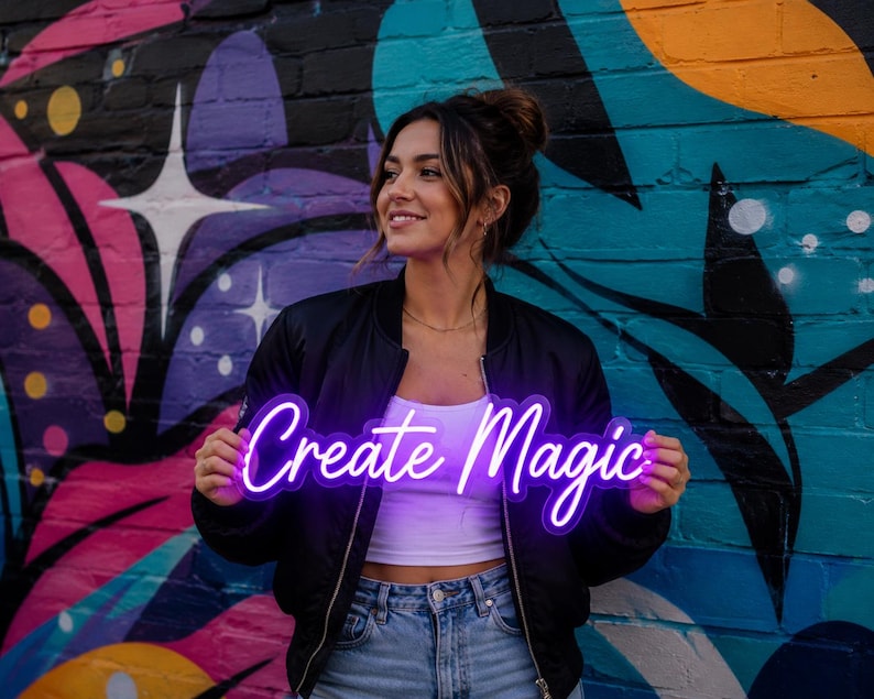 May include: A bright purple neon sign featuring the text Create Magic in a cursive script font. The sign is illuminated and emits a vibrant glow, showcasing the words clearly against the dark background of the black bomber jacket.