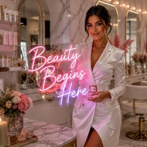 May include: A rectangular clear acrylic neon sign featuring the pink glowing text Beauty Begins Here in a cursive script font. The sign is held in front of a person wearing a white satin blazer dress with a matching belt and a gold buckle.