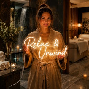 May include: A glowing neon sign with the text Relax & Unwind in a cursive script font. The sign is constructed from clear acrylic backing and bright white neon tubing, emitting a soft light that illuminates the surrounding area.