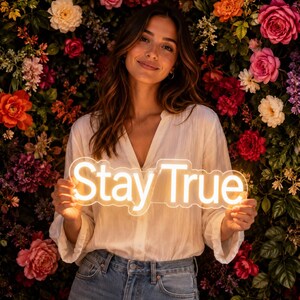 May include: A white neon sign featuring the text Stay True in a glowing script font. The sign is constructed with a clear acrylic backing that follows the contour of the letters, emitting a warm light that illuminates the surrounding area.