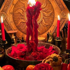 K&ouml;nnte beinhalten: Eine rituelle Szene mit einer brennenden roten Kerze in Form von zwei sich umarmenden Figuren, vor einem Holz Hintergrund. Eine Schale mit roten Rosenbl&auml;ttern und Fr&uuml;chten ist von brennenden roten Kerzen umgeben.