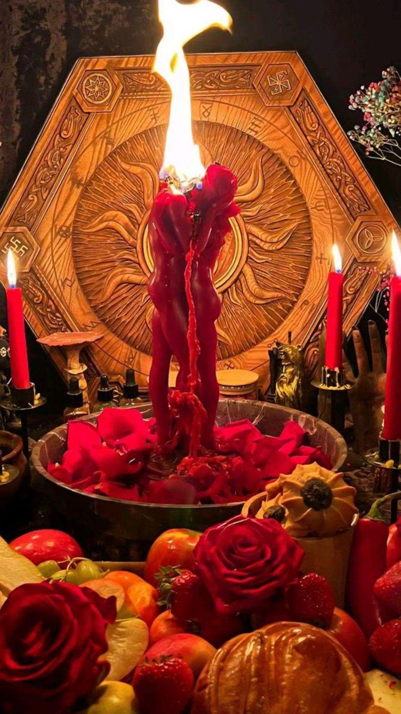 K&ouml;nnte beinhalten: Eine rituelle Szene mit einer brennenden roten Kerze in Form von zwei sich umarmenden Figuren, vor einem Holz Hintergrund. Eine Schale mit roten Rosenbl&auml;ttern und Fr&uuml;chten ist von brennenden roten Kerzen umgeben.
