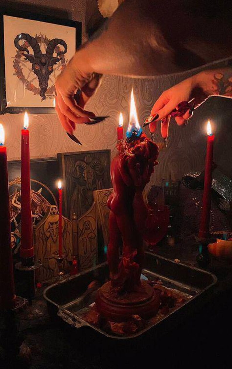 May include: A red candle sculpture of two figures embracing, with a blue flame. Several red candles are lit around the sculpture. A framed image of a horned figure is in the background, along with other occult-themed items.