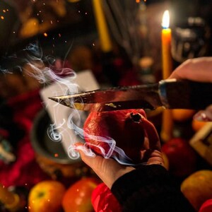May include: A hand holding a red pomegranate being cut with a knife. Smoke rises from the pomegranate. Other fruits and a lit candle are in the background. The scene is set on a red surface.