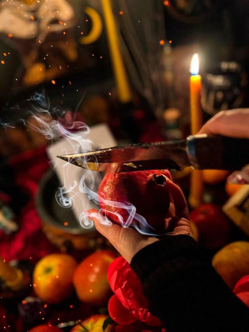 May include: A hand holding a red pomegranate being cut with a knife. Smoke rises from the pomegranate. Other fruits and a lit candle are in the background. The scene is set on a red surface.